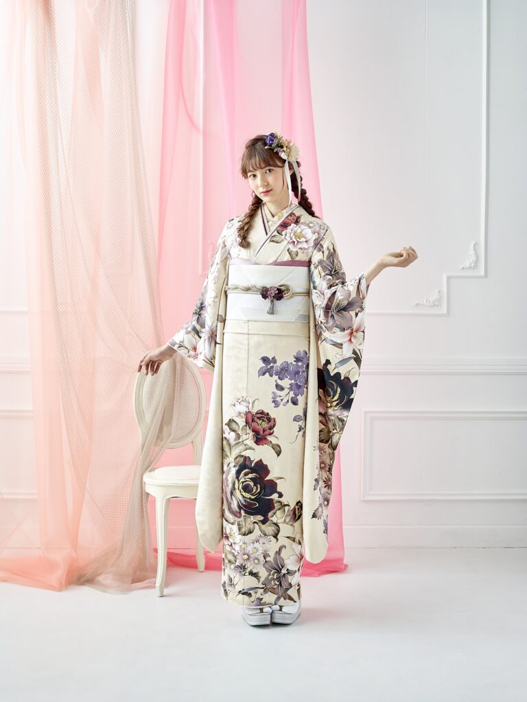 クリーム色の花柄の着物を着た女性が室内に立っている。紫と黒の花があしらわれた三つ編みの髪を結い、白い椅子の横で優雅なポーズをとっている。背景には白い壁と透け感のあるピンクのカーテンが映えている。.