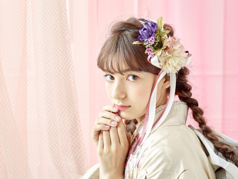 茶色の髪を編み込み、花柄のヘアアクセサリーをつけた若い女性が、クリーム色の着物を着て、両手を顔の近くで組んでカメラを見つめています。背景には淡いピンクと白のドレープ生地があしらわれ、優しく優雅な雰囲気を醸し出しています。.