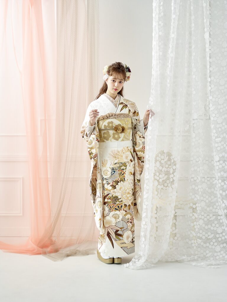 室内には、白と金に繊細な花柄が施された華やかな着物を着た若い女性が立っています。花の髪飾りをつけ、透け感のあるレースのカーテンを掲げています。背景には、淡いピンクと白のドレープと、モールディングが施された白い壁が描かれています。.