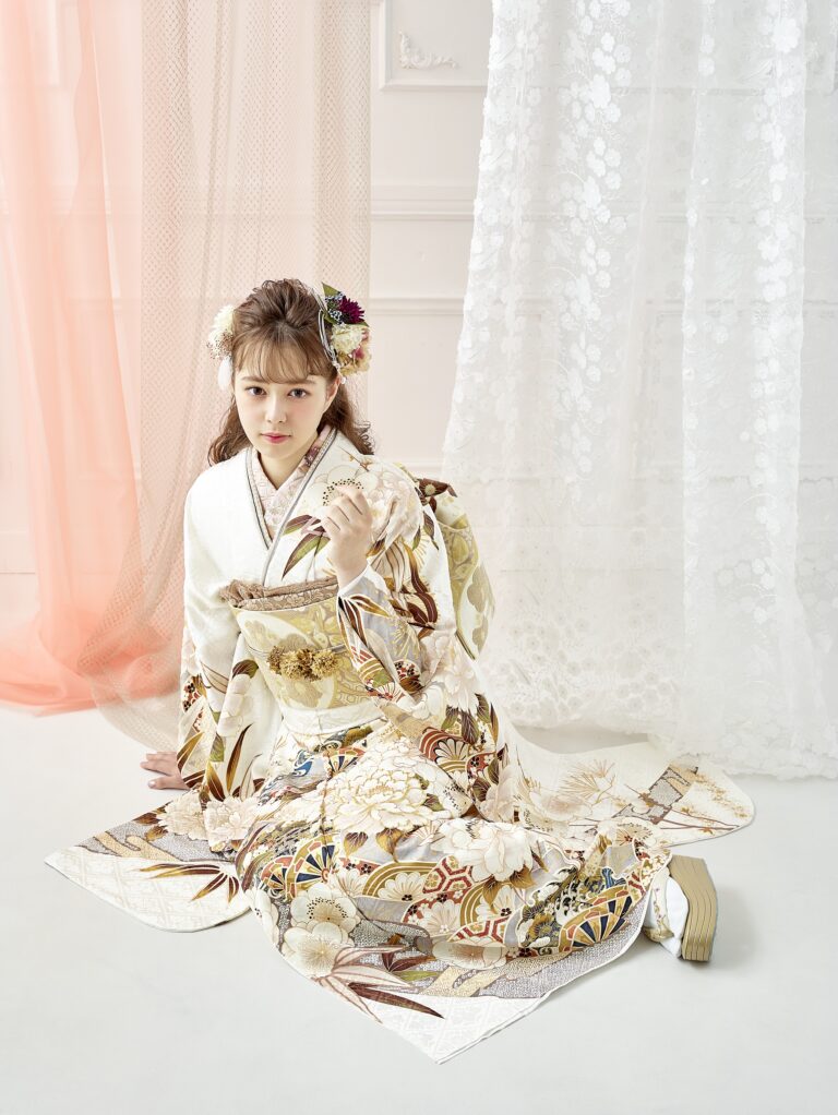 金、茶色、花柄があしらわれた華やかな白い着物を着た若い女性が、床にひざまずいている。髪には花を飾り、小さな装飾品を持っている。明るい背景には、ピンクと白の柔らかな薄手のカーテンがかかっている。.