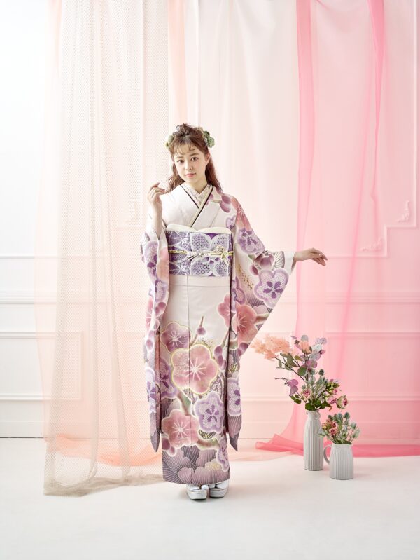 白とラベンダーの花柄の着物に帯を締めた若い女性が立っている。髪には花を飾り、片手を顔の近くに持っていく。彼女の背後には、透け感のあるピンクのカーテンと、床に置かれたパステルカラーの花が飾られた花瓶が、柔らかく優雅な雰囲気を醸し出している。.