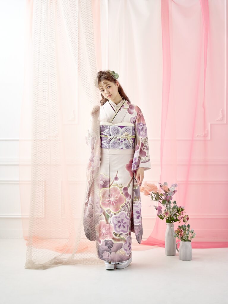 淡い紫の花柄の着物を着た若い女性が、透け感のあるピンクとベージュのドレープがかかった明るい部屋に立っています。髪には花を飾り、白いサンダルを履いています。彼女の隣には、パステルカラーの花が入った花瓶が置かれています。その光景は、柔らかな光に包まれ、優雅な雰囲気を醸し出しています。.