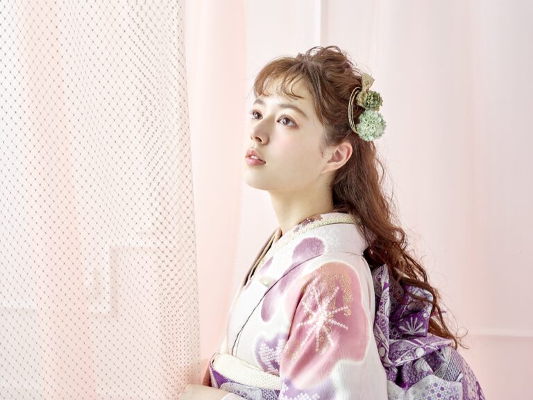 ピンクと白の花柄の着物に紫の帯を締めた若い女性が、室内に立って上を見つめている。ウェーブヘアに白と緑のヘアアクセサリーを添えている。柔らかな光が彼女の顔と、網戸のカーテンがかかったパステルピンクと白の背景を照らしている。.