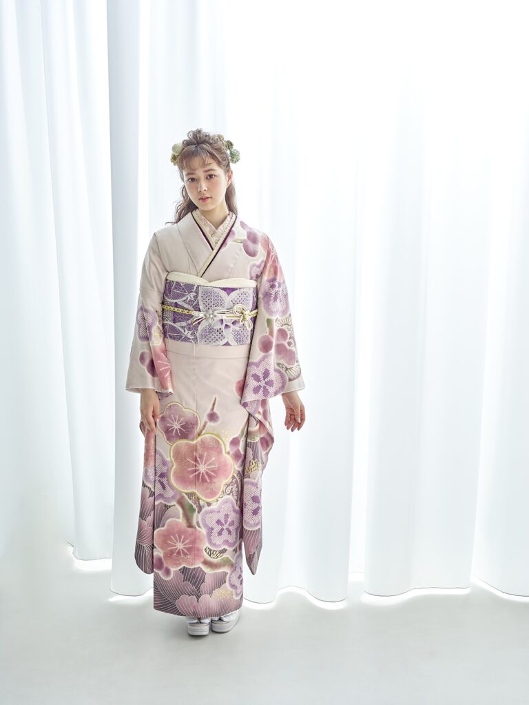 淡いピンク色の着物に紫とピンクの花柄をあしらった若い女性が、白い幕の前に立っている。髪には小花を飾り、穏やかな表情で両手を優しく脇に当てている。柔らかな照明が差し込んでいる。.