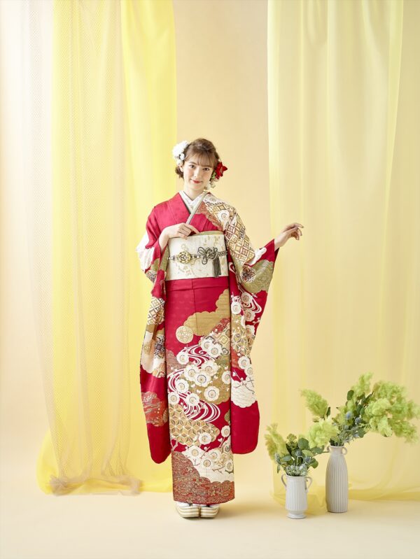若い女性が、赤、金、白の花柄の着物を着た柔らかな黄色のカーテンの前に立っています。髪には花を飾り、片方の袖を差し出しています。彼女の隣には、緑の葉が飾られた白い花瓶が2つ床に置かれています。背景は薄黄色です。.