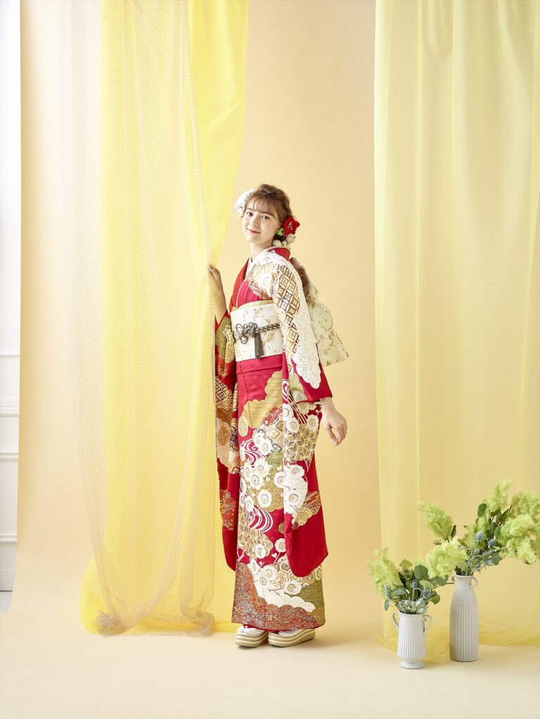 紅白の花柄の着物を着た若い女性が、微笑みながら黄色の透けた幕を手に立っています。伝統的な髪飾りと草履を履いています。背景には柔らかな黄色のカーテンが敷かれ、床には緑の花が入った白い花瓶が2つ置かれています。.