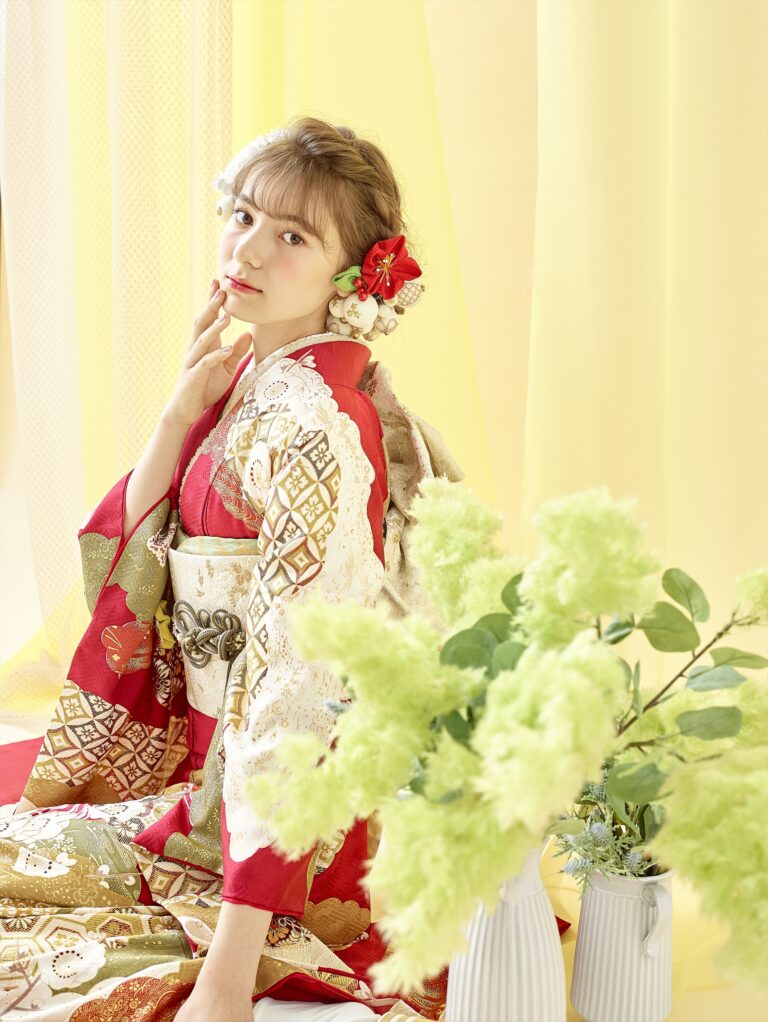 赤と金の花柄の伝統的な着物を着た若い女性が、優雅にカメラを見つめています。髪には赤い花も含めた花が飾られ、柔らかな黄色のカーテンと白い花瓶に飾られた緑の花に囲まれています。.