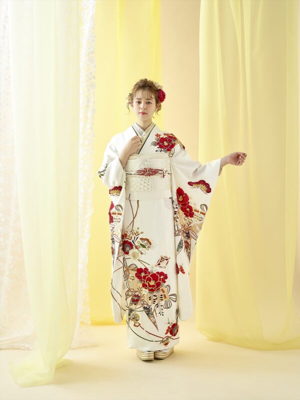 淡い黄色のカーテンを背に、赤と金の花柄が入った白い着物を着た若い女性が立っている。明るい髪をアップにし、赤い花飾りを添え、白い草履を履いている。右腕を上げ、着物の袖を見せている。.