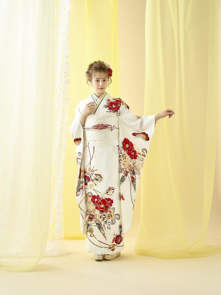 透け感のある黄色のカーテンの前に、赤と金の花柄があしらわれた白い着物を着た女性が立っている。巻き毛に赤い花を添え、片腕を優雅に挙げてポーズをとっている。背景は柔らかな光に照らされ、優雅な雰囲気を醸し出している。.