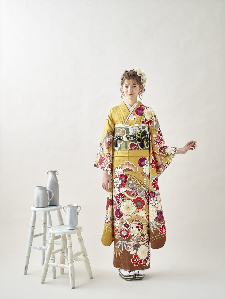 シンプルな背景に、黄色の花柄の着物に黒と金の帯を締めた若い女性が立っています。短い巻き毛には白い花が飾られています。彼女の傍らには、白いスツールと水差しが巧みに積み重ねられ、装飾的な雰囲気を醸し出しています。.