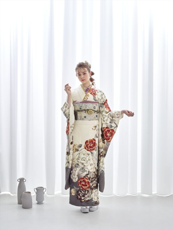 クリーム色の着物を着た女性が、白い幕の前に立っています。着物には紅白の大きな花模様が描かれています。華やかな髪飾りと白い草履を身につけています。彼女の左側の床には、陶器の水差しと花瓶が並べられています。.
