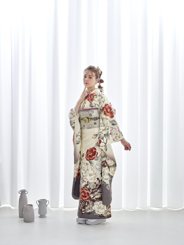 クリーム色の生地に赤と灰色の花柄があしらわれた3073（複製）の着物を着た若い女性が、白いカーテンの前に立っている。彼女の髪は飾りのヘアピースでまとめられており、彼女の横の床には灰色と白の陶器の水差しが置かれている。.