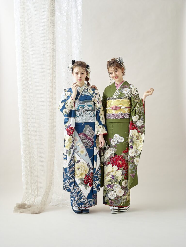 二人の女性が、色鮮やかな花柄の着物を着て並んで立っています。左の女性は青い着物に白と赤の花が、右の女性は緑の着物に黄と赤の花が描かれています。二人とも花の髪飾りをしており、落ち着いた表情で、明るい背景を背景にポーズをとっています。.