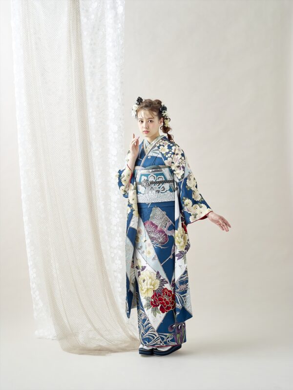 透け感のある白いカーテンの前に、青と白の花柄の着物を着た若い女性が立っている。花で飾られた髪をセットし、片手を顔の近くに、もう片手を横に伸ばして、優しくカメラを見つめている。.
