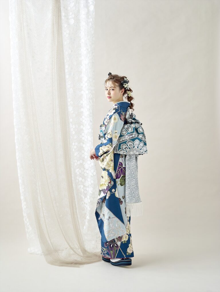 透け感のある質感のあるカーテンの前に、青と白の花柄の着物に華やかな帯を締めた若い女性が横顔で立っている。髪には花のアクセサリーを添え、明るくミニマルなスタジオの雰囲気の中で、優しくカメラを見つめている。.
