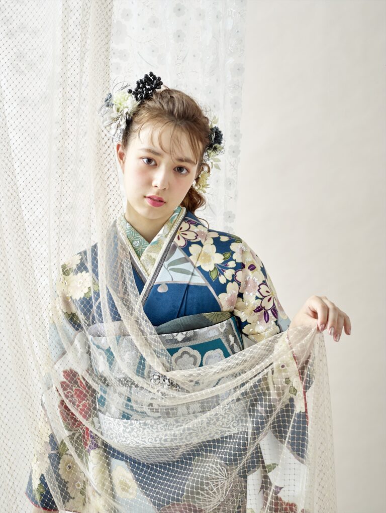 青い花柄の着物を着た若い女性が、透け感のあるレースの布を持って立っています。髪には花飾りが飾られ、カメラを優しく見つめています。背景は明るく、彼女の伝統的な和装の優雅さと細部までこだわった美しさを引き立てています。.