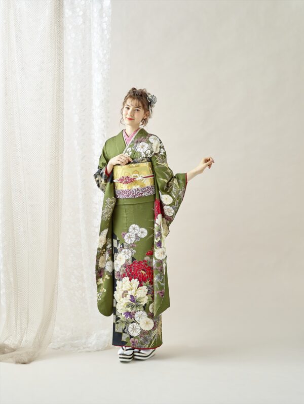 緑の花柄の着物に金色の帯、白い足袋、白黒の草履を履いた女性が室内に立っている。薄茶色の髪をアップヘアにし、ヘアアクセサリーをつけて、淡い背景を背景に片手を挙げて優しく微笑んでいる。.