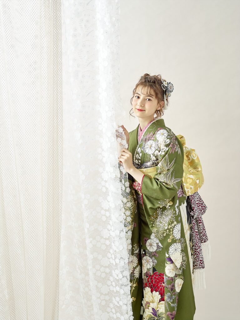 緑の花柄の着物に黄色の華やかな帯を締めた若い女性が、透け感のある白い模様のカーテンのそばに立っていて、カーテンを優しく分けている。髪にはヘアピンと花飾りが飾られ、明るく落ち着いた背景の中で優しく微笑んでいる。.