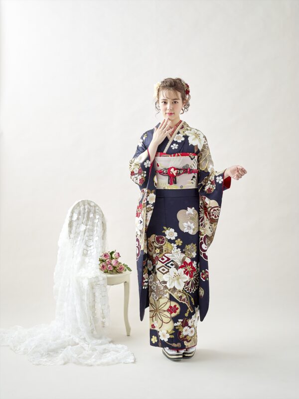 紺とクリーム色に赤、金、白の模様が入った花柄の着物を着た若い女性が、白い背景に立っている。彼女は片方の袖をまくり上げ、唇に触れている。彼女の隣には、白いレースがかけられた椅子があり、ピンクと赤の小さな花束が置かれている。.