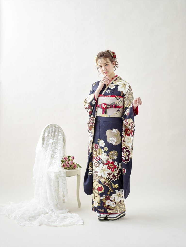 紺地に花柄の装飾が施された着物を着た女性が、レースがかけられた白い椅子の横に立っています。彼女はピンクと赤の小さな花束を手に持っています。背景には、伝統的な草履と髪飾りを身につけ、柔らかなニュートラルカラーが映えています。.