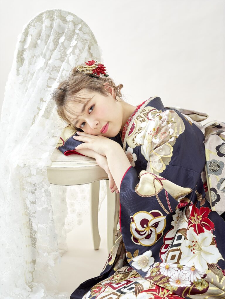 金、赤、白の模様が入った濃い花柄の着物を着た若い女性が、レースがかけられた白い椅子に頭と腕をもたせかけている。淡いニュートラルな背景を背景に、赤い花のアクセサリーで髪をまとめ、カメラを見つめている。.