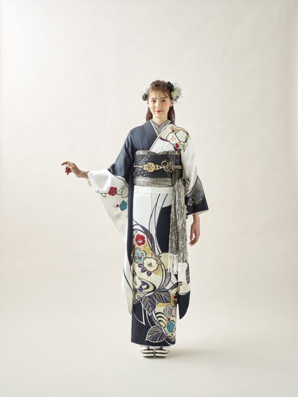スタジオには、花柄の伝統的な白黒の着物と華やかな帯を締めた若い女性が立っています。髪には花を飾り、明るい背景に片腕を少し横に伸ばしています。.
