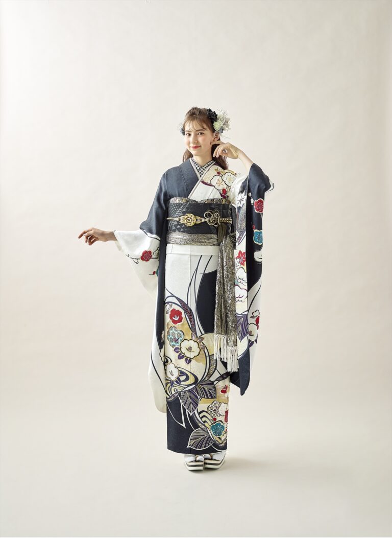 スタジオには、白黒の花柄のフォーマルな着物「3113（複製）」を身につけた女性が立っている。背景には、無地で淡い色合いの帯、白い花の髪飾り、足袋、草履が添えられている。彼女は優しく微笑んでいる。.