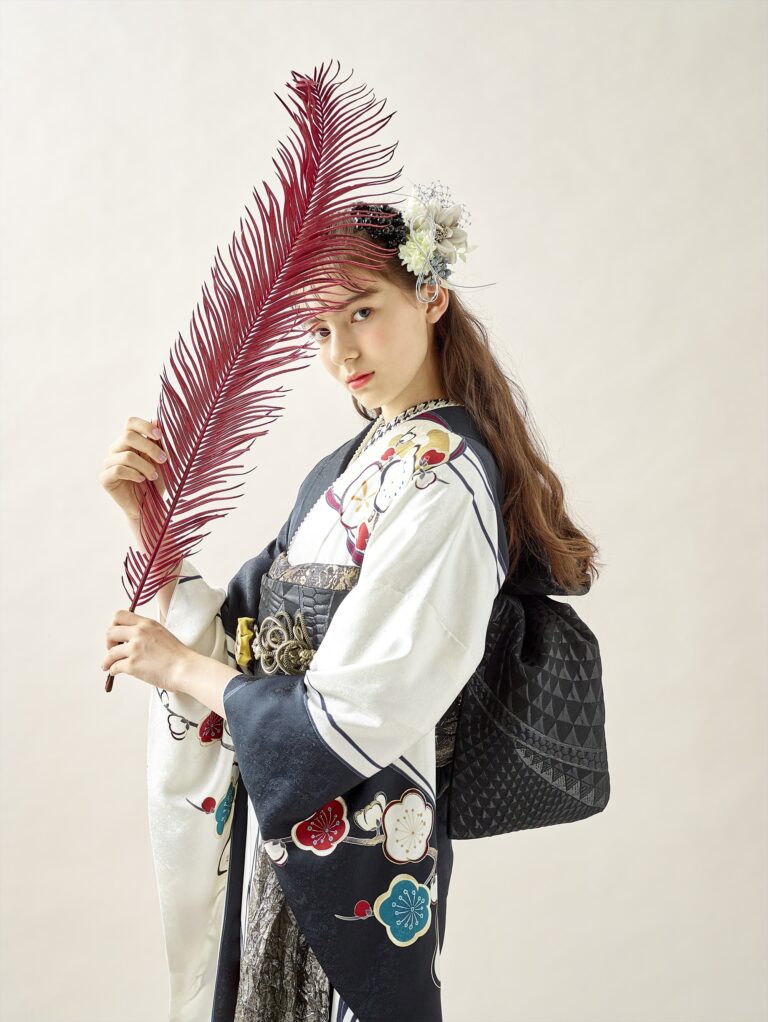 柄物の着物を着た若い女性が、大きな赤いヤシの葉を前に掲げている。彼女は商品番号3113（複製）を着用し、白い花をつけた長い茶色の髪をしており、明るい背景を背にカメラに向かっている。.