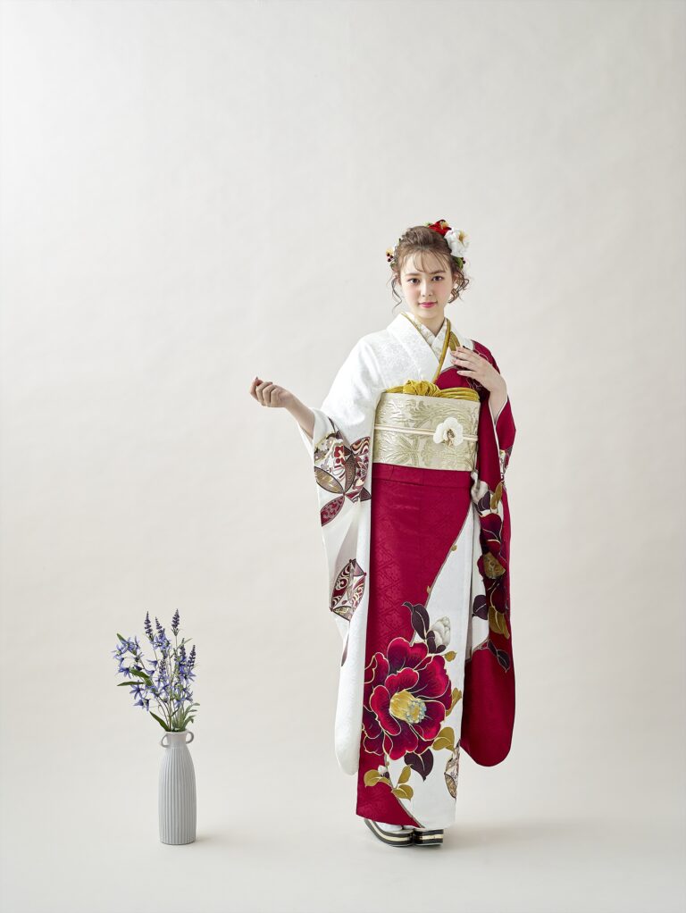 スタジオに立つ女性は、大胆な花柄があしらわれた白と深紅の着物、華やかな帯、そして花の髪飾りを身に着けている。彼女は、無地の明るい背景を背に、紫と緑の花が入った白い花瓶の横で優雅なポーズをとっている。.