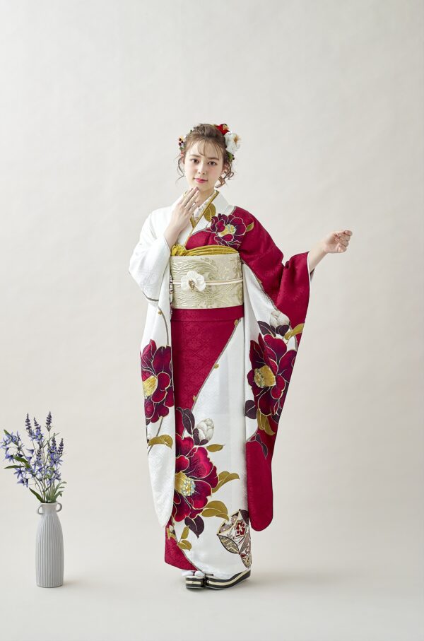 白と深紅の花柄の着物に金の帯を締めた女性が、淡い背景に優雅に立っている。髪には花を飾り、片手を口元に上げている。彼女の左側の床には、紫色の花が飾られた背の高い花瓶が置かれている。.