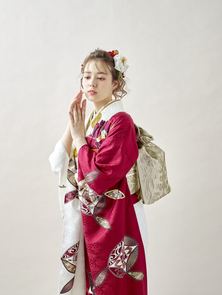 紅白の模様が入った日本の伝統的な着物を着た若い女性が横顔で立っている。ウェーブヘアに花を飾り、金色の帯を後ろで結んでいる。両手を顔の近くで優しく合わせ、考え込むように横を向いている。.