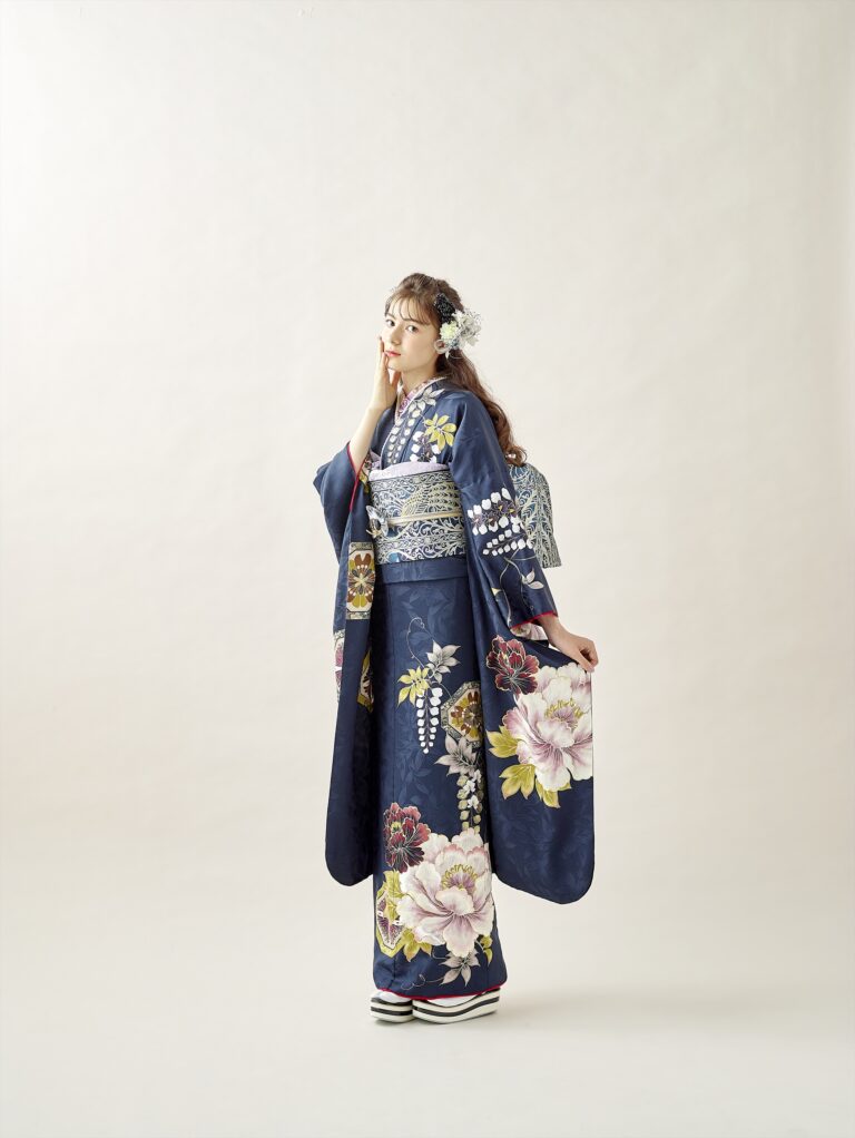 明るい背景に、ピンク、白、黄色の大きな花柄があしらわれた紺色の着物を着た若い女性が立っています。花の髪飾りと伝統的な草履を身につけ、片手を顔の近くに上げて優雅なポーズをとっています。.