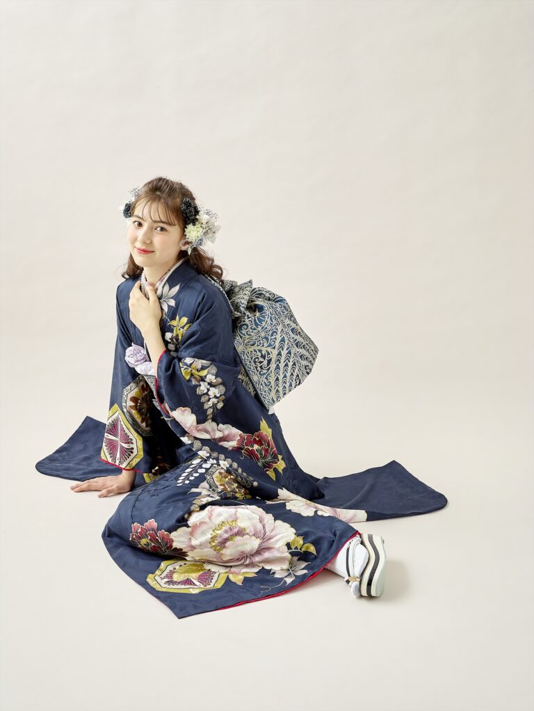 紺色の花柄の着物と白い足袋を履いた若い女性が、明るい背景にひざまずいている。花柄の髪飾りをつけ、柄物の帯を腰に下げ、カメラに向かって優しく微笑んでいる。.