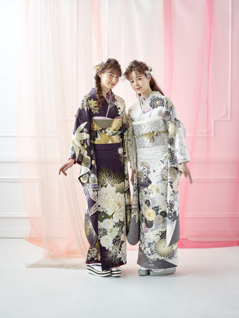 華やかな着物を着た二人の女性が、ピンクと白の透け感のあるカーテンの前に並んで立っています。一人は濃い紫色に金と白の花柄が入った着物、もう一人は薄い灰色に銀と金の模様が入った着物を着ています。二人とも花柄の髪飾りをつけています。.