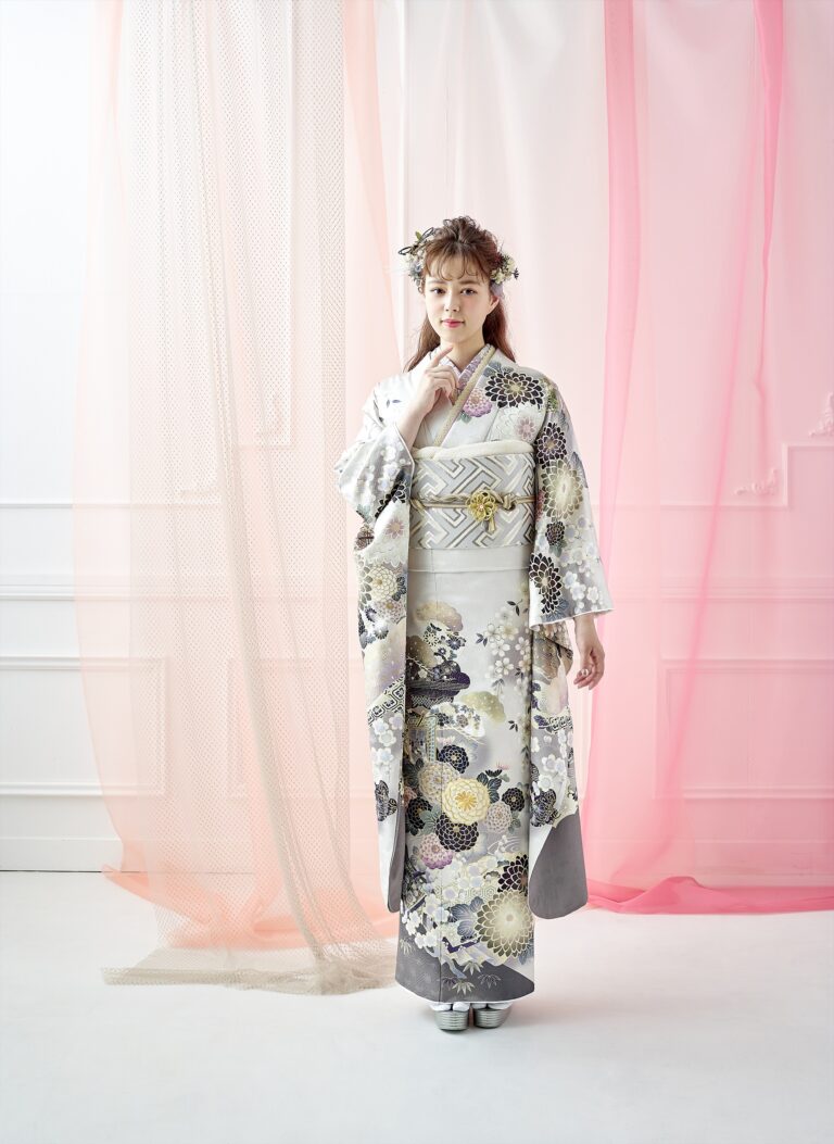 白いスタジオに、銀と白の繊細な花柄のフォーマルな着物を着た女性が立っている。髪には花のヘッドピースを着け、片手を顔に近づけてポーズをとっている。彼女の背後には、ピンクと白の透け感のある布のドレープが垂れ下がっている。.