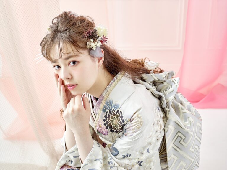 淡い花柄の着物を着た若い女性が室内でひざまずき、二本の指で優しく頬に触れている。ウェーブのかかった髪には小さな花が飾られている。背景には淡いピンクとベージュのドレープが描かれ、優雅で繊細な雰囲気を醸し出している。.