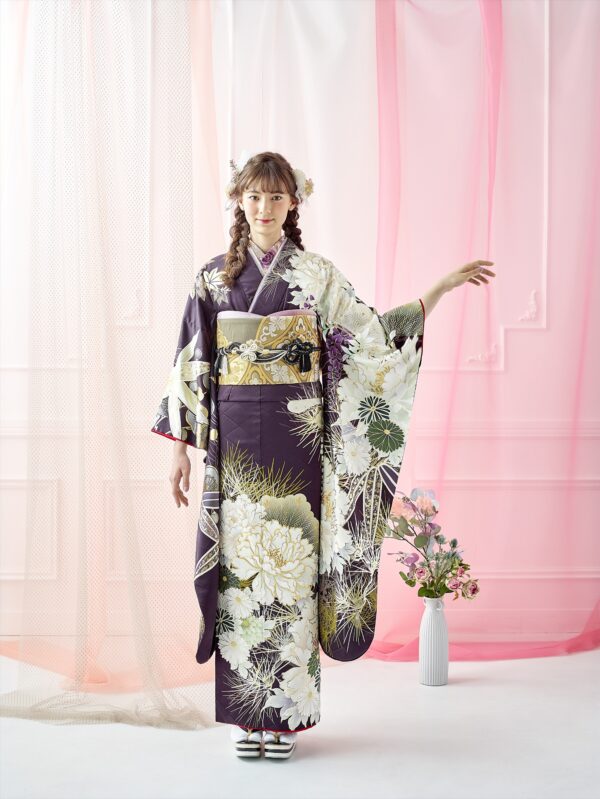 若い女性が、白と金の模様が入った濃い色の花柄の着物を着て、室内に立っています。片腕を上げてポーズをとっています。髪には花が飾られています。彼女の後ろには、白とピンクの透け感のあるカーテンがかかっており、床には背の高い花瓶が置かれています。.