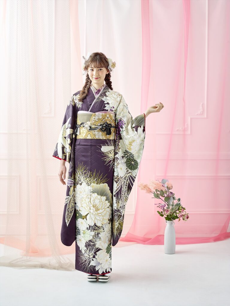 白と金の花柄が入った黒い着物を着た若い女性がスタジオに立っている。編み込んだ髪に花を添え、片腕を軽く上げている。彼女の背後には、ピンクの透け感のあるカーテンとパステルカラーの花が飾られた白い花瓶が、その光景を彩っている。.