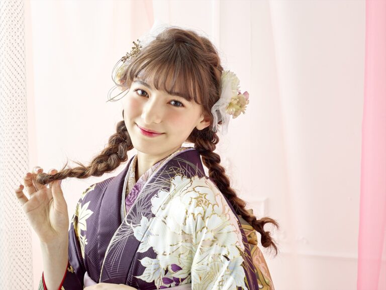 白と金の模様があしらわれた紫の花柄の着物を着た若い女性が、優しく微笑んでいます。茶色の長い髪を二つ編みにし、白い花を飾っています。柔らかなパステルピンクの背景が、優しく伝統的な雰囲気を醸し出しています。.