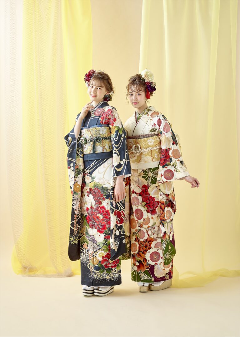 二人の女性が、伝統的な帯と髪飾りをつけた色鮮やかな花柄の着物を着て並んで立っています。一人は青い着物に大きな赤い花が描かれ、もう一人は白い着物に赤と緑の模様が描かれています。二人とも柔らかな黄色の布のカーテンの前で優しく微笑みながらポーズをとっています。.