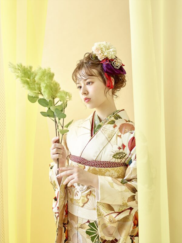 クリーム色の花柄の着物を着た若い女性が、薄い黄色のカーテンのそばに立っており、緑の葉のついた枝を手に持っています。彼女の髪には大きな花と装飾品が飾られています。背景は黄色で、柔らかく優雅な雰囲気を醸し出しています。.