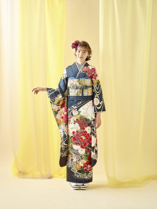 黄色い背景を背に、微笑む女性が立っている。色鮮やかな花柄の紺色の着物に金色の帯、白い足袋に草履を履いている。短いウェーブヘアには大きな赤い花が飾られ、背景には透けるような黄色のカーテンがかかっている。.
