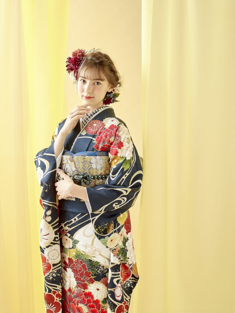 淡い黄色の背景に、赤、白、金の花柄があしらわれた紺色の伝統的な着物を着た若い女性が立っています。髪には赤い花を添え、軽く顎に触れながら、優しくカメラを見つめています。.