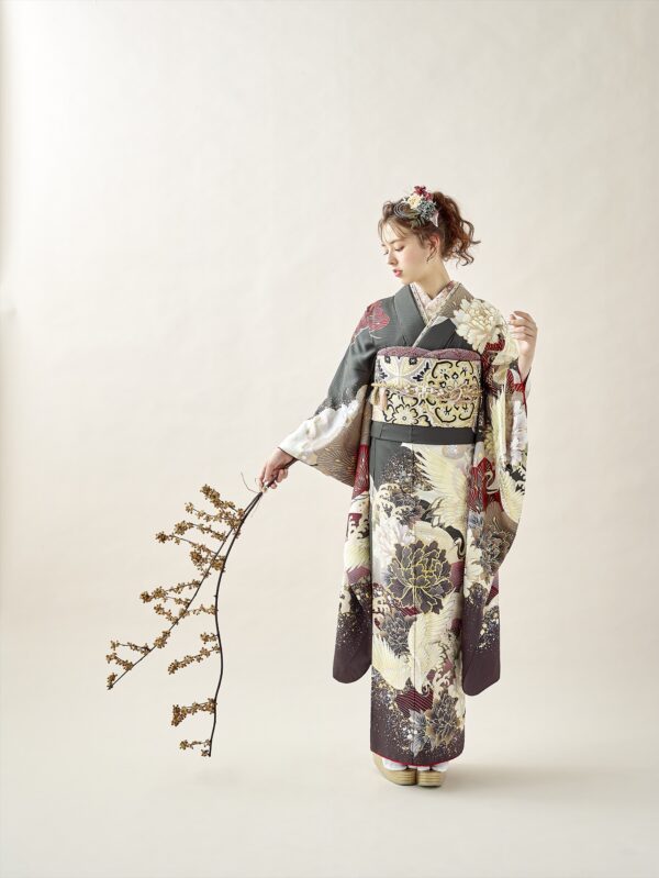 金、黒、象牙、赤の模様があしらわれた華やかな着物を着た女性が、小さな花が咲いた枝を持って立っています。彼女は花で飾られた豪華なアップヘアで、柔らかなベージュの背景に映える枝を見下ろしています。.