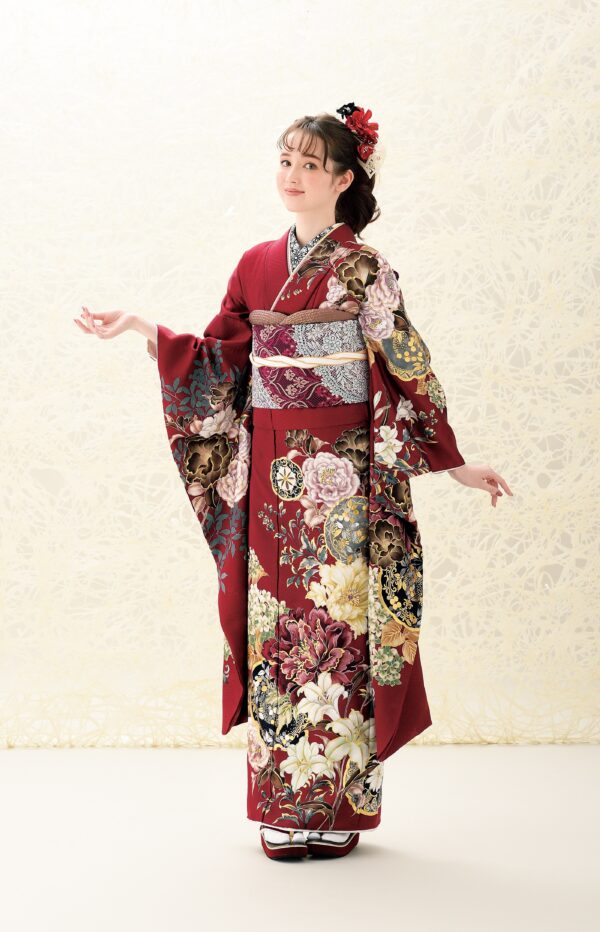 クリーム色の背景に、金、白、ピンクのアクセントが入った深紅の花柄の着物をまとった若い女性が立っています。赤い花で飾られた髪をセットし、腕を軽く伸ばし、白い足袋と草履を履いて優雅なポーズをとっています。.