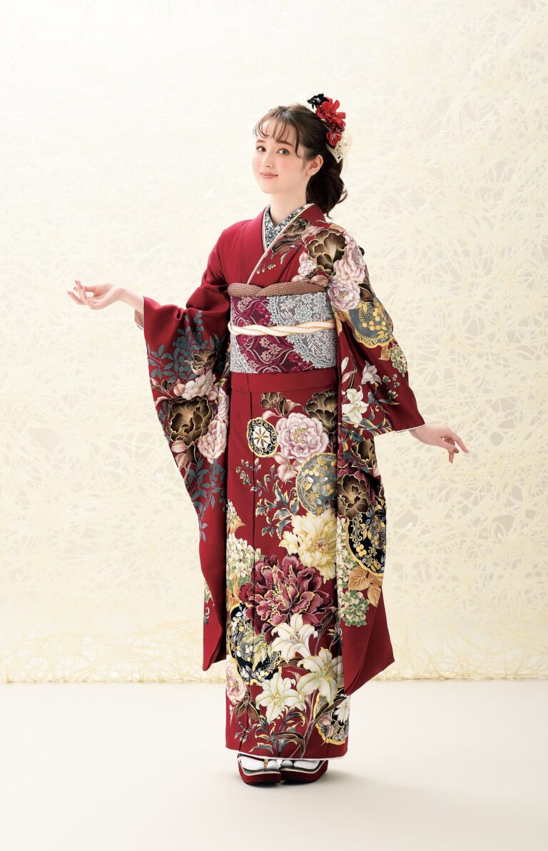 クリーム色の背景に、金、白、ピンクのアクセントが入った深紅の花柄の着物をまとった若い女性が立っています。赤い花で飾られた髪をセットし、腕を軽く伸ばし、白い足袋と草履を履いて優雅なポーズをとっています。.