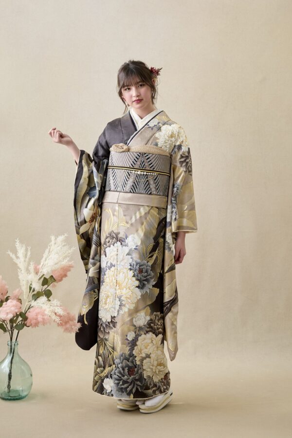 ベージュの背景に、金とグレーの華やかな花柄の着物に模様の帯を締めた若い女性が立っています。髪には花を添え、表情は穏やかです。彼女の左側には、羽毛のような質感の白と淡いピンクの花が飾られたガラスの花瓶が置かれています。.