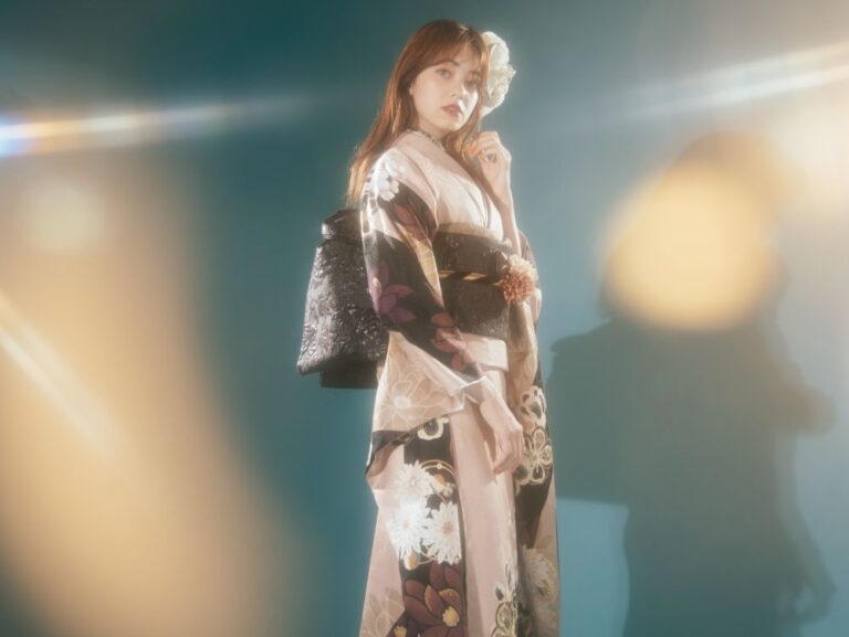 青緑色の背景に、柄のある着物を着た女性がポーズをとっています。着物には白、紫、ベージュの花柄が描かれています。彼女は華やかな黒の帯を締め、白い花を顔に添えています。明るい光のフレアが、柔らかく幻想的な雰囲気を醸し出しています。.