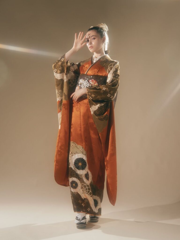 ベージュの背景に、オレンジと茶色の伝統的な模様の着物を着た人物が立っています。精巧な鬘をつけ、片手を顔の近くに持っています。柔らかな照明が柔らかな輝きを放ち、白黒の履物を履いています。この光景は、優雅さと文化遺産の雰囲気を醸し出しています。.