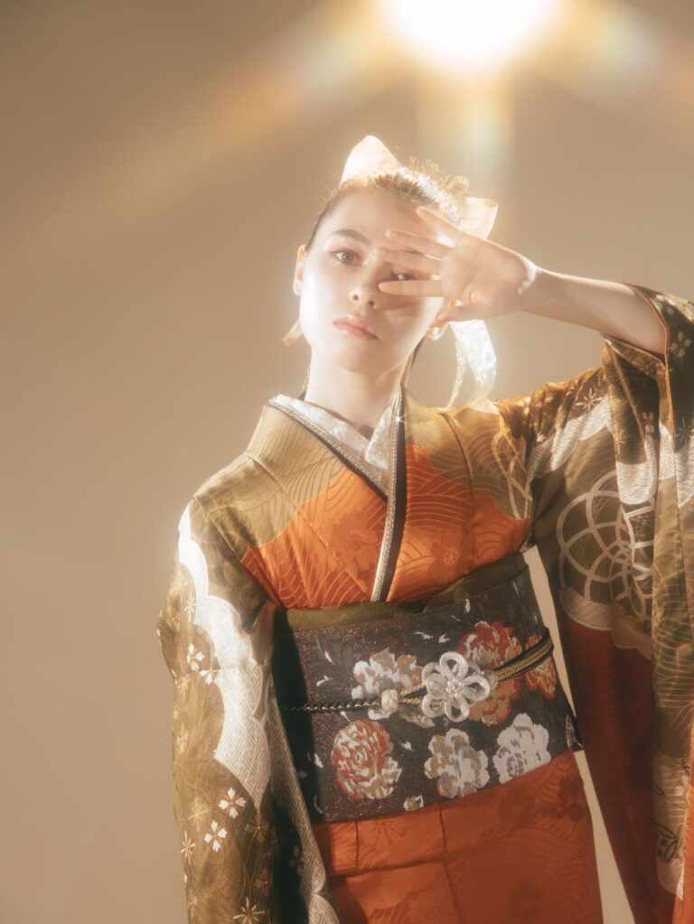 オレンジと茶色の伝統的な着物を着た人物が、片手で顔を覆いながらポーズをとっている。着物には花柄が描かれ、刺繍が施された濃い色の帯が締められている。柔らかく温かみのある照明が柔らかな光を放ち、髪に結んだリボンが美しく映えている。.